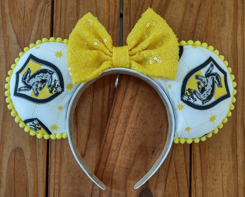 May include: Yellow and white fabric headband with a yellow bow and a white fabric patch with a black and yellow crest featuring a rabbit and the words "Hufflepuff" and "House".