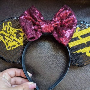 May include: Black sequined Minnie Mouse ears with a burgundy bow and gold glitter lettering that says "Hufflepuff" on one ear and a Hogwarts crest on the other.