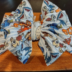 May include: A blue and white fabric bow with a Finding Nemo theme. The bow features characters like Dory, Nemo, and Marlin, as well as sea turtles and other sea creatures. The bow is tied with a white ribbon.