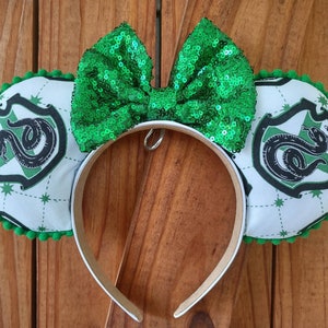 May include: Green and white mouse ears with a green sequin bow. The ears feature the Hogwarts house crest for Slytherin with a green snake and the words "Slytherin" in a white banner.