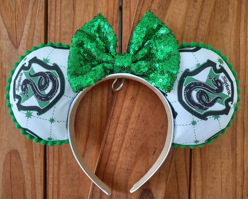 May include: Green and white mouse ears with a green sequin bow. The ears feature the Hogwarts house crest for Slytherin with a green snake and the words "Slytherin" in a white banner.