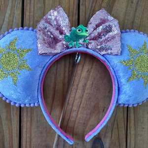 May include: Purple Minnie Mouse ears with a gold glitter sun design on each ear. A pink sequined bow with a green chameleon charm is attached to the headband.