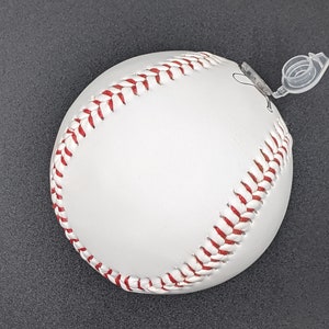 May include: White baseball with red stitching. The ball has a clear plastic tab attached to it.