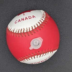 May include: A red and white baseball with the word "CANADA" printed on it. The ball has a white valve for inflating.