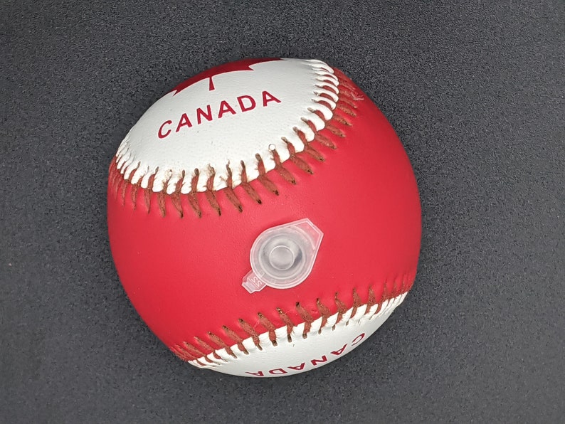May include: A red and white baseball with the word "CANADA" printed on it. The ball has a white valve for inflating.