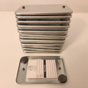 May include: A stack of 15 silver metal boxes with a single box open and a white paper logbook inside. The logbook has the text "Geocaching Game Play" and "Name/Date" printed on it.