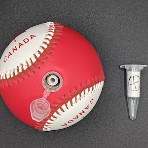 May include: A red and white baseball with the word "CANADA" printed on it. The baseball has a clear plastic valve on the side. A small clear plastic tube with a white label and a black cross symbol is next to the baseball.