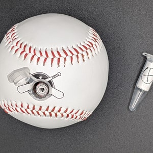 May include: White baseball with red stitching and a black and white logo of two crossed bats. A small clear plastic tube with a white label and a black cross is in the background.