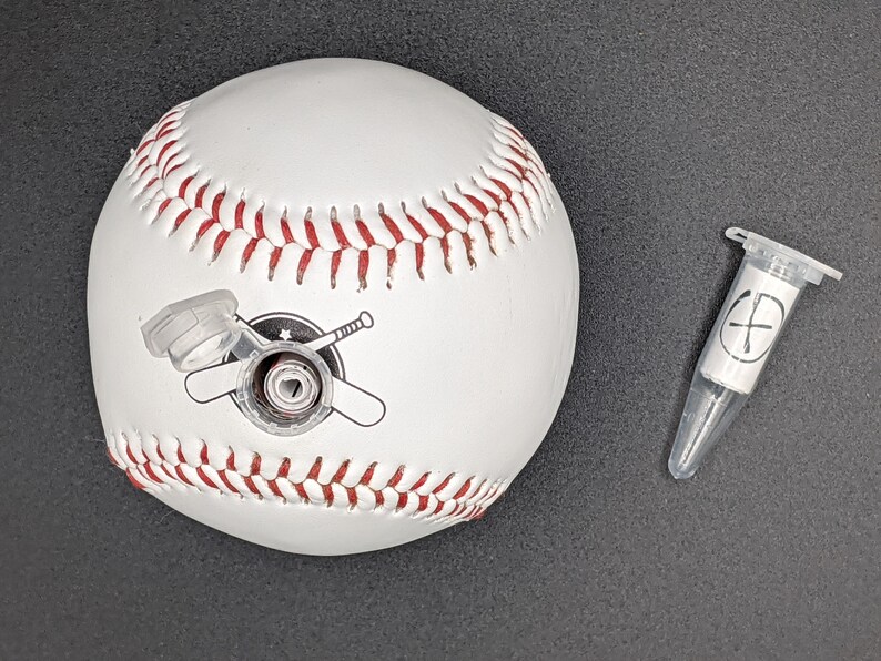 May include: White baseball with red stitching and a black and white logo of two crossed bats. A small clear plastic tube with a white label and a black cross is in the background.