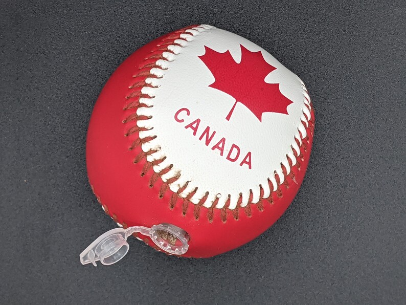 May include: A red and white baseball with a Canadian flag and the word "CANADA" printed on it. The baseball has a white valve on the side.