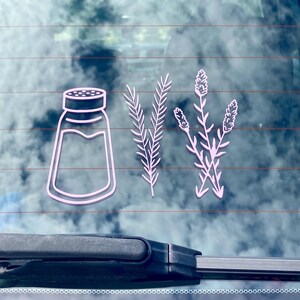 May include: A white car window decal with a salt shaker, rosemary sprigs, and lavender flowers. The design is in a minimalist style.