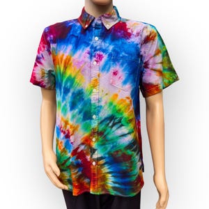May include: Short-sleeved button-down shirt with a vibrant tie-dye pattern in shades of blue, purple, yellow, and orange. The shirt has a chest pocket and a classic collar. The shirt is displayed on a mannequin.