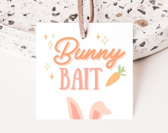 Puede incluir: Una etiqueta blanca con el texto "Bunny Bait" en letras naranja y rosa. La etiqueta tiene una pequeña zanahoria naranja y dos orejas de conejo rosas en la parte inferior. La etiqueta está atada a una cuerda marrón.