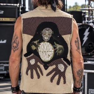 May include: A beige sleeveless vest featuring a large back patch with a skull graphic, the text "CARNIVAL MADNESS," and two brown plaid fabric hands with white floral accents. The vest has a brown plaid yoke and a collar, worn over dark jeans.