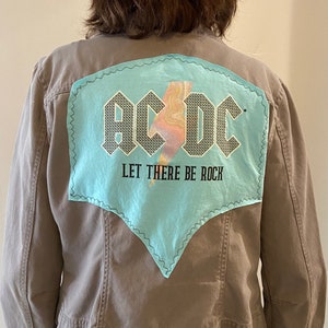 May include: A gray jacket with a light blue patch featuring the band name "AC/DC" and a lightning bolt graphic. The text "LET THERE BE ROCK" is below the band name. The patch is outlined with a decorative stitch.