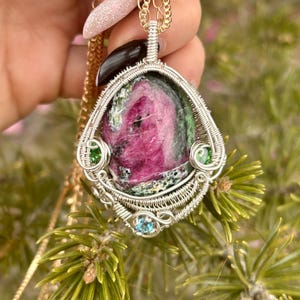Puede incluir: Un colgante envuelto en alambre plateado con una piedra preciosa rosa y verde con piedras preciosas verdes y azules más pequeñas. El colgante cuelga de una cadena de oro.