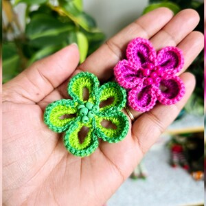 Op de afbeelding: Twee handgemaakte gehaakte bloemapplicaties. Eén is groen met een lichtergroen centrum en kralen, de andere is roze met een donkerderroze centrum en kralen. De bloemen worden in een hand gehouden, met groen gebladerte op de achtergrond.