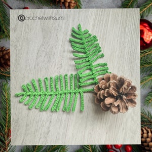 Może przedstawiać: Dwa zielone liście paproci wykonane na szydełku i brązowy szyszkowy na jasnym drewnianym tle. Tekst "@crochetwithsumi" znajduje się w lewym górnym rogu obrazu.