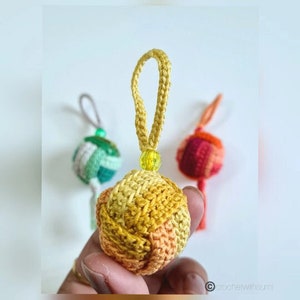 Puede incluir: Un adorno de bola de crochet en tonos amarillos, naranjas y rosas. El adorno tiene un lazo para colgar y una pequeña cuenta en la parte superior.