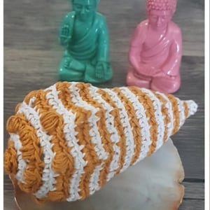 May include: A crocheted seashell in white and orange stripes sits on a natural stone coaster. Two small, colorful Buddha statues are in the background.