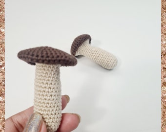 Op de afbeelding: Twee gehaakte paddenstoelen, een bruin en wit, de andere wit met een bruine dop. De paddenstoelen zijn klein en gemaakt van een getextureerd garen.