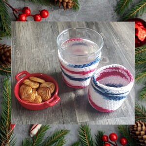 Puede incluir: Dos vasos, uno lleno de agua, y un plato pequeño con galletas. Los vasos están decorados con fundas de punto a rayas rojas, blancas y azules. Decoraciones festivas navideñas rodean los objetos.