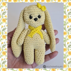 May include: A crocheted yellow bunny with long ears, wearing a yellow scarf and a yellow bow. The bunny is holding a small yellow flower.