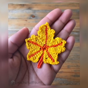 May include: A crochet maple leaf applique featuring bright yellow yarn for the leaf body and vibrant orange yarn for the central veins and stem. The text crochetwithsumi is printed in the bottom left corner of the image.