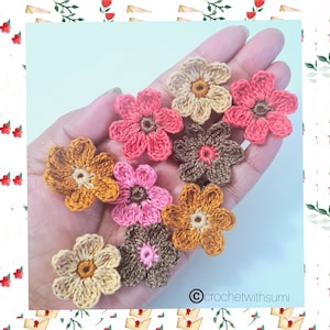 May include: A collection of nine crocheted flowers in various colors, including pink, orange, brown, and cream. The flowers are arranged in a hand, with the copyright symbol and the text "crochetwithsumi" visible in the bottom right corner.