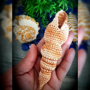 Puede incluir: Una concha de mar de ganchillo en tonos beige y marrón. La concha tiene forma de espiral y una superficie texturizada.