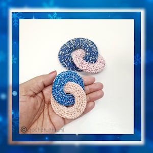 Puede incluir: Dos anillos de ganchillo, uno azul y otro rosa, están entrelazados. Los anillos están hechos con un hilo texturizado y tienen un acabado ligeramente brillante.