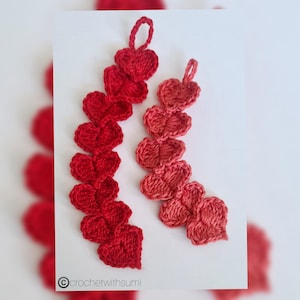 May include: Two crocheted heart garlands, one in red and one in a gradient of pink and red. The garlands are made of individual crocheted hearts that are connected together.