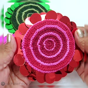 May include: Close-up of handmade crochet flower embellishments. The central flower is a spiral design in shades of red and pink, surrounded by red sequins. Another flower with green and dark green crochet and green sequins is visible in the background. The image is held by hands.