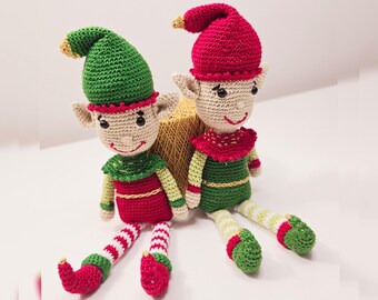 May include: Two crocheted elf ornaments with green and red hats, green and red outfits, and white and green striped legs. The elves are sitting on a white surface.