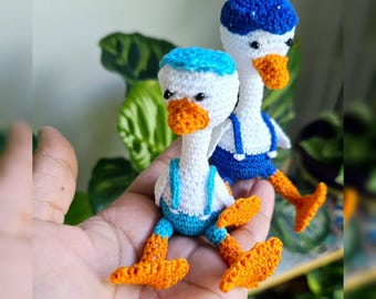 May include: Two crocheted duck plushies wearing blue overalls and blue and white hats. The ducks are sitting on a hand.