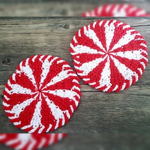 May include: Two red and white crocheted pot holders shaped like candy canes. The pot holders are on a wooden surface.