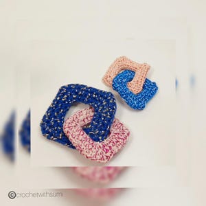 Puede incluir: Tres cuadrados de ganchillo hechos a mano en azul, rosa y azul con detalles de hilo metálico dorado. Los cuadrados están entrelazados, creando un diseño único. La imagen está sobre un fondo blanco. El texto "© crochetwithsumi" está en la esquina inferior izquierda.