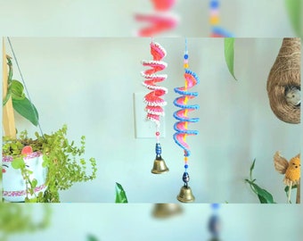 May include: Two crocheted spiral wind chimes with bells hanging from them. The wind chimes are pink and white, and blue and yellow. The wind chimes are hanging from a white wall.