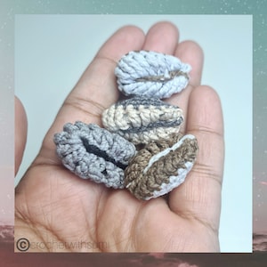 May include: Five crocheted coffee beans in various shades of brown, gray, and white. The beans are held in a hand.