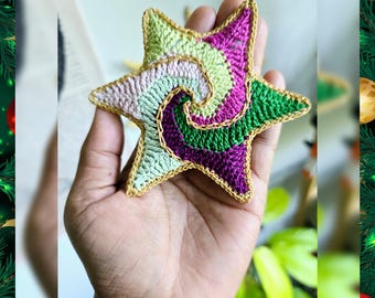 May include: A crocheted star ornament with a spiral design in shades of pink, green, and gold. The star is held in a hand.