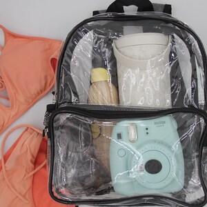 May include: A clear plastic backpack with a black zipper and black trim. The backpack is filled with various items, including a blue instant camera, a clear water bottle, a white container, and sunglasses.