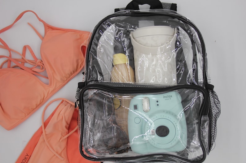 May include: A clear plastic backpack with a black zipper and black trim. The backpack is filled with various items, including a blue instant camera, a clear water bottle, a white container, and sunglasses.