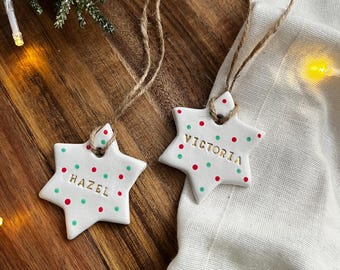 Könnte beinhalten: Zwei weiße sternförmige Weihnachtsschmuckstücke mit roten und grünen Punkten und den Namen "HAZEL" und "VICTORIA" in goldfarbenen Buchstaben. Jedes Schmuckstück hat eine Aufhängung aus Juteschnur. Die Schmuckstücke liegen auf einer Holzoberfläche.