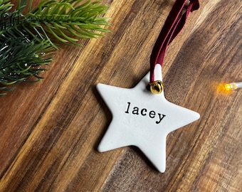 May include: White star-shaped ornament with the name "Lacey" printed in black. It has a burgundy ribbon and a small gold bell. The ornament is on a wooden surface with a sprig of greenery.