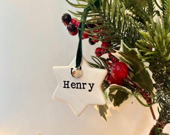 May include: A white star-shaped Christmas ornament with the name "Henry" printed in black. The ornament hangs from a green ribbon and a small silver bell. The ornament is surrounded by green pine needles and red berries.