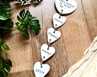 May include: A vertical hanging decoration featuring five white heart-shaped plaques connected by twine. The top heart reads "This nanny is loved by..." while the four hearts below are inscribed with the names "Oliver," "Thomas," "George," and "Albie."