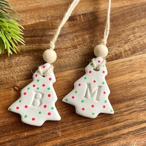 Puede incluir: Dos adornos navideños de cerámica blanca con forma de árbol, con lunares rojos y verdes. Cada árbol tiene una letra, "B" y "M", y una cuenta de madera en la parte superior. Cuelgan de una cuerda de hilo natural.