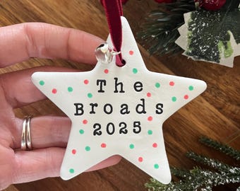 Könnte beinhalten: Weißer sternförmiger Weihnachtsschmuck mit roten und grünen Tupfen. Der Text "The Broads 2025" ist schwarz gedruckt. Ein rotes Band und eine silberne Glocke sind zum Aufhängen befestigt. Festliche Weihnachtsdekoration.
