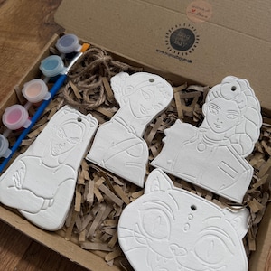 May include: A craft kit with four unpainted ceramic ornaments in a cardboard box. The ornaments include three female figures and a cat face. The kit also contains paint pots and a paintbrush, ready for creative painting.
