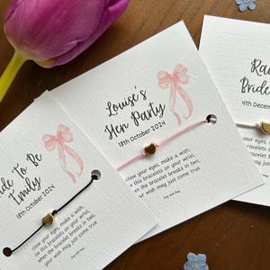 May include: Two white cards with pink ribbons and gold heart charms attached to black and pink string bracelets. The cards have text that reads "Bride to Be Emily 18th October 2024" and "Louise's Hen Party 18th October 2024". The text on the cards also includes the phrase "close your eyes, make a wish, tie this bracelets on your wrist, when the bracelet breaks in two, your wish may just come true".